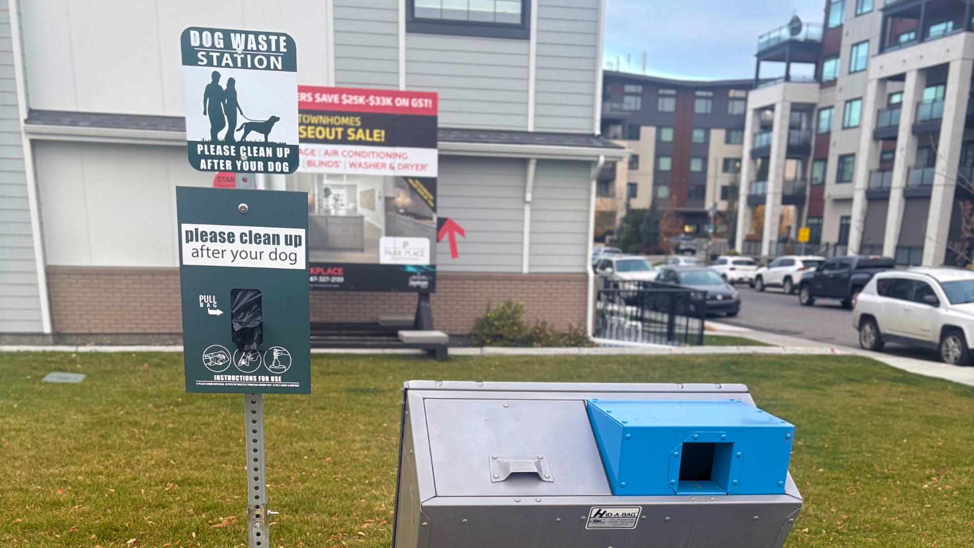 A new green dog waste station with a bag dispenser, next to a metal waste receptacle on the lawn at Central Green